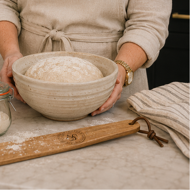 A woman wearing glasses and a beige apron stands in a kitchen holding a large bowl of flour. On the counter in front of her are a kraft paper bag of flour with a visible 'Sourdough' label, a small glass jar of flour, a wooden rolling pin, and a striped cloth.